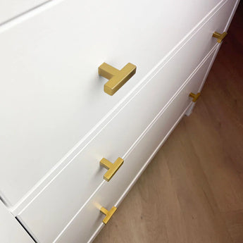 Gros plan d'une commode blanche avec des boutons Vigio Gold de yessential.fr sur trois tiroirs, chacun décoré de poignées dorées modernes en forme de T, sur un parquet marron clair.