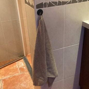 Une serviette grise est suspendue à un crochet Rudro Black yessential.fr, fixé au mur carrelé de la salle de bain près de la douche, au-dessus du carrelage marron et beige.