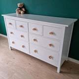 La commode Bambino Heart de yessential.fr dispose de huit tiroirs blancs avec boutons cœur, nuage, étoile et lune. Un ours en peluche marron sur le dessus ajoute du charme à cette chambre d'enfant, contre un mur vert et un parquet en bois clair.