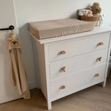 Une commode blanche avec des poignées yessential.fr Bambino Star en forme d'étoiles, de nuages ​​et de lunes, surmontée d'un matelas à langer beige et d'un panier à jouets en peluche. Une serviette à capuche beige avec un visage d'animal est suspendue à proximité - idéale pour n'importe quel thème de boutons de chambre d'enfant ou de chambre d'enfant.