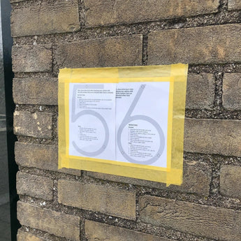 Une feuille de numéro de maison Amato White de 12,5 cm de yessential.fr, montrant les chiffres 5 et 6 ainsi que des instructions textuelles, est collée sur un mur de briques avec du ruban adhésif jaune, suggérant qu'elle fait partie d'un guide ou d'un affichage séquentiel.