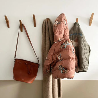 Un sac à main en cuir marron, une écharpe beige, une doudoune rose de dessin animé et un manteau matelassé vert sont accrochés au porte-manteau Fabio Oak yessential.fr contre un mur bicolore blanc et beige.