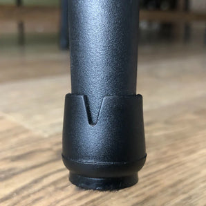 Gros plan d'un embout de pied noir Paco Set Ø 25-29mm de yessential.fr sur un pied de meuble, reposant sur un parquet en bois. Le capuchon présente une encoche en forme de V et une surface lisse et légèrement brillante pour protéger le sol.