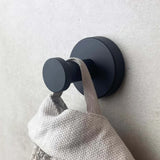 Un crochet mural rond noir mat de l'ensemble de WC Xavo Black de yessential.fr maintient une serviette ou un sac texturé gris clair contre un mur de couleur claire.