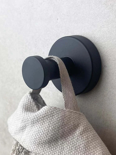Un crochet mural rond noir mat de l'ensemble de WC Xavo Black de yessential.fr maintient une serviette ou un sac texturé gris clair contre un mur de couleur claire.