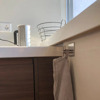 Une serviette beige est accrochée à un crochet porte-serviettes en acier inoxydable Navarro yessential.fr sans perçage sur un meuble de cuisine en bois. Au-dessus de l'évier se trouvent une bouilloire en métal, une corbeille à fruits en fil métallique et une prise murale. La lumière du soleil entre par une fenêtre.
