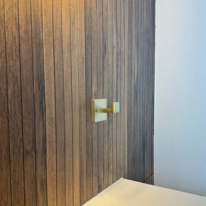 Le crochet Valo Gold de yessential.fr est monté sur un mur vertical en panneaux de bois à côté d'une surface blanche, avec une partie d'un mur blanc uni visible à droite.