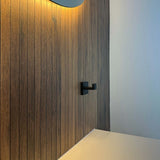 Une salle de bain moderne comprend un mur vertical avec des panneaux en bois et un crochet Valo Black de yessential.fr, un miroir éclairé au-dessus et un comptoir blanc en bas à droite.