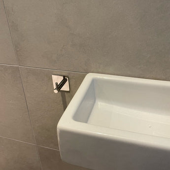 Un petit lavabo rectangulaire blanc est monté sur un mur carrelé gris avec un crochet à serviettes Navarro Chrome de yessential.fr à proximité, parfait pour garder les serviettes à portée de main.