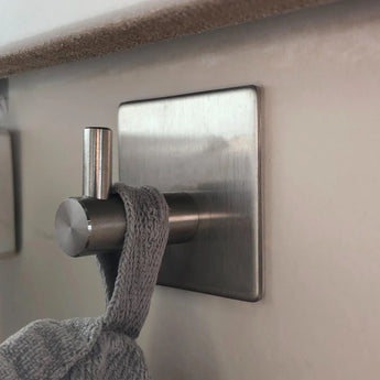 Un gros plan d'un crochet à serviette en acier inoxydable Navarro yessential.fr sans perçage tenant une serviette en tricot gris contre un mur de couleur claire.