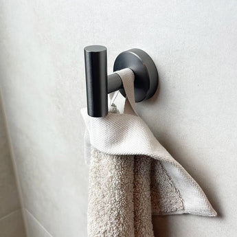 L'ensemble WC Nando Gunmetal de yessential.fr comprend une serviette grise pliée et un chiffon blanc sur un élégant crochet mural en métal noir, élégamment montés sur un mur légèrement carrelé - idéal pour moderniser la salle de bain.