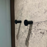 Quatre crochets Carreno noirs de yessential.fr sont montés sur un mur texturé avec un motif en pierre. Les crochets projettent des ombres douces vers la droite et un verre ou une surface réfléchissante apparaît partiellement sur le côté gauche de l'image.