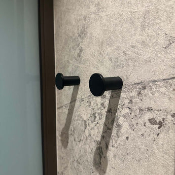 Quatre crochets Carreno noirs de yessential.fr sont montés sur un mur texturé avec un motif en pierre. Les crochets projettent des ombres douces vers la droite et un verre ou une surface réfléchissante apparaît partiellement sur le côté gauche de l'image.