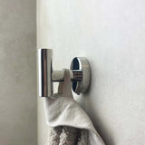 Un gros plan montre le crochet Rodrigo Chrome yessential.fr, un accessoire de salle de bain cylindrique moderne, solidement monté et tenant une serviette texturée beige contre un mur de couleur claire.