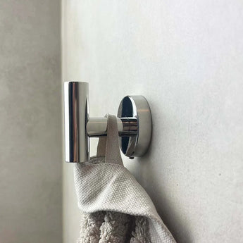Un gros plan montre le crochet Rodrigo Chrome yessential.fr, un accessoire de salle de bain cylindrique moderne, solidement monté et tenant une serviette texturée beige contre un mur de couleur claire.
