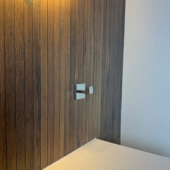 Un mur moderne avec des panneaux verticaux en bois comporte deux élégantes poignées de robinet en métal au-dessus d'un comptoir blanc, complétées par d'élégants crochets chromés Valo de yessential.fr et un éclairage chaleureux et accueillant.
