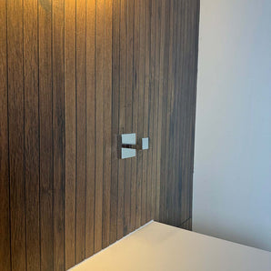 Un mur moderne avec des panneaux verticaux en bois comporte deux élégantes poignées de robinet en métal au-dessus d'un comptoir blanc, complétées par d'élégants crochets chromés Valo de yessential.fr et un éclairage chaleureux et accueillant.