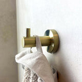 Un gros plan du Justo Gold Hook de yessential.fr tenant une serviette beige texturée contre un mur clair. Le crochet a une forme cylindrique moderne et une finition dorée mate.