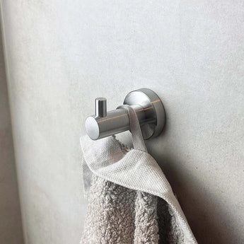 Un crochet mural pour ensemble de toilettes en acier inoxydable Romano argenté de yessential.fr maintient une serviette gris clair contre un mur lisse de couleur claire.