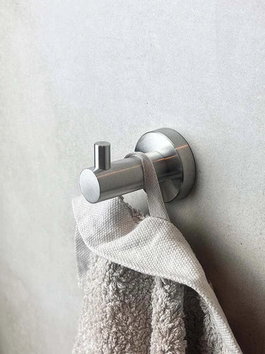 Un crochet mural pour ensemble de toilettes en acier inoxydable Romano argenté de yessential.fr maintient une serviette gris clair contre un mur lisse de couleur claire.
