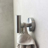 L'ensemble de toilettes Ferro en acier inoxydable de yessential.fr comprend un crochet mural élégant et cylindrique en métal argenté au design minimaliste, qui maintient solidement une serviette texturée contre un mur de couleur claire.