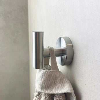 Un gros plan de l'ensemble de toilettes Corrado en acier inoxydable de yessential.fr, avec le crochet mural rond argenté monté sur un mur clair, avec une serviette beige soigneusement enroulée dessus.