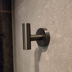 Le crochet Rodrigo Gunmetal de yessential.fr présente un design métallique élégant et cylindrique. Monté sur un mur carrelé gris, il ajoute une touche moderne et une teinte subtile – idéal pour n'importe quelle salle de bain.