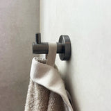 L'ensemble de toilettes Quirino Gunmetal de yessential.fr composé d'une murale moderne en crochet et cylindrique en métal avec une boucle avec texture serviette en beige, montée sur un mur - idéal pour un bain minimaliste.