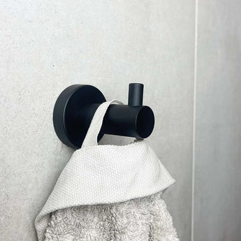 L'ensemble de toilette Donato Black de yessential.fr comprend un crochet mural noir mat qui maintient une serviette texturée blanche contre des carreaux gris clair, ajoutant un style moderne à vos accessoires de salle de bain.