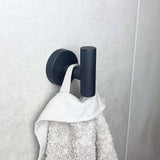 Le crochet noir Rodrigo de yessential.fr maintient solidement une serviette blanche avec une boucle de suspension contre un mur légèrement carrelé, ajoutant une élégance moderne au décor de votre salle de bain.