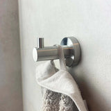 Une serviette grise avec une boucle en tissu est suspendue à un crochet argenté pour ensemble de toilettes en acier inoxydable Marino de yessential.fr, monté sur un mur clair. Le design cylindrique et la petite cheville s'adaptent à tous les accessoires de salle de bain modernes.