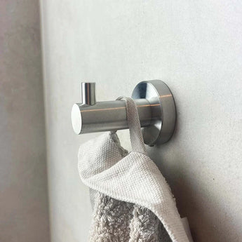 Une serviette grise avec une boucle en tissu est suspendue à un crochet argenté pour ensemble de toilettes en acier inoxydable Marino de yessential.fr, monté sur un mur clair. Le design cylindrique et la petite cheville s'adaptent à tous les accessoires de salle de bain modernes.