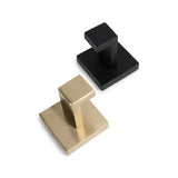 Le crochet Valo Gold de yessential.fr et un crochet à serviette carré noir mat sont disposés sur une surface blanche. Chaque crochet en acier inoxydable a un design géométrique et angulaire avec une base et un bras rectangulaires.