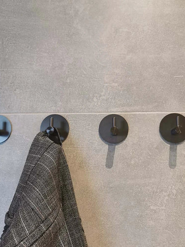 Quatre crochets ronds Rudro Gunmetal de yessential.fr sur un mur carrelé gris, avec un vêtement à carreaux gris foncé sur le deuxième crochet. Les autres sont vides – idéal comme crochet porte-serviettes sans perçage pour les salles de bains modernes.