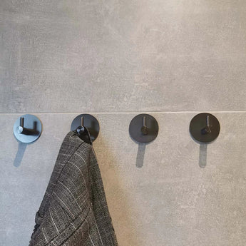Quatre crochets Delo Gunmetal WC Set de yessential.fr sont montés sur un mur carrelé gris. Un manteau à motifs gris est suspendu au deuxième crochet, tandis que les autres sont vides, parfait pour une configuration d'accessoires de toilette modernes sans perçage.