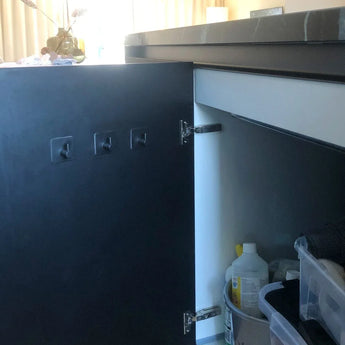La porte d'un placard de cuisine est ouverte et montre des produits de nettoyage. Quatre crochets Navarro Gunmetal de yessential.fr sont fixés à l'intérieur de la porte, actuellement inutilisés. La lumière du soleil provenant d'une fenêtre éclaire l'arrière-plan.