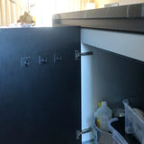 La porte d'une armoire de cuisine est ouverte, révélant des produits de nettoyage et des conteneurs. Quatre crochets Navarro Black de yessential.fr sont fixés à l'intérieur de la porte, mais ces crochets pratiques ne contiennent actuellement rien.