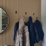 Quatre manteaux sont suspendus aux 10 crochets Huso Black yessential.fr fixés à un mur vertical à lattes de bois. À gauche, un miroir rond au cadre noir reflète en partie l’espace soigné et moderne.