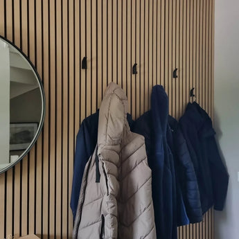 Quatre manteaux sont suspendus aux 10 crochets Huso Black yessential.fr fixés à un mur vertical à lattes de bois. À gauche, un miroir rond au cadre noir reflète en partie l’espace soigné et moderne.