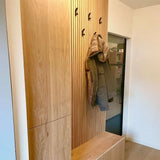 Un porte-manteau moderne en bois avec 10 crochets Huso Black de yessential.fr contient un manteau vert. Des placards intégrés et un banc se trouvent en dessous, tandis qu'une porte vitrée en arrière-plan reflète l'espace intérieur.