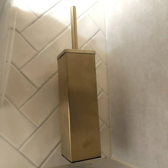 Le porte-brosse de toilette Alessino Gold de yessential.fr, avec une poignée dorée assortie, se trouve dans le coin contre des murs carrelés en diagonale blanche.