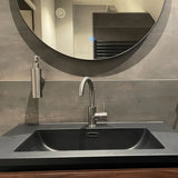 L'Alberto en acier inoxydable de yessential.fr comprend un lavabo noir mat, un robinet argenté, un distributeur de savon mural et un grand miroir rond au-dessus qui reflète l'intérieur de la salle de bain.