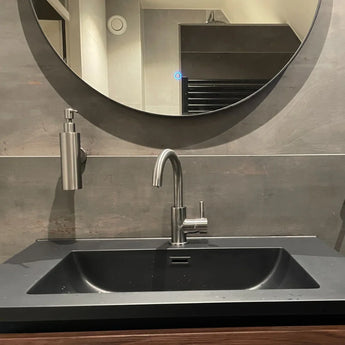 L'Alberto en acier inoxydable de yessential.fr comprend un lavabo noir mat, un robinet argenté, un distributeur de savon mural et un grand miroir rond au-dessus qui reflète l'intérieur de la salle de bain.