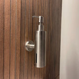 La pompe à savon Alberto en acier inoxydable de yessential.fr, avec un design cylindrique mince et une pompe sur le dessus, est fixée au mur sur un mur vertical en bois avec des lattes étroites et égales.
