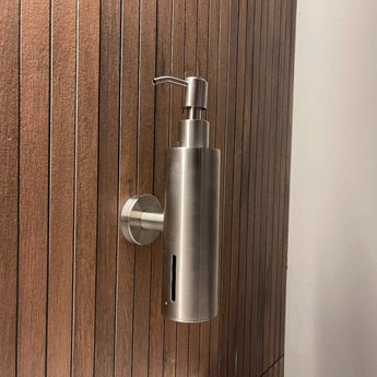 La pompe à savon Alberto en acier inoxydable de yessential.fr, avec un design cylindrique mince et une pompe sur le dessus, est fixée au mur sur un mur vertical en bois avec des lattes étroites et égales.