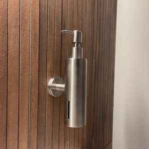 La pompe à savon Alberto en acier inoxydable de yessential.fr, avec un design cylindrique mince et une pompe sur le dessus, est fixée au mur sur un mur vertical en bois avec des lattes étroites et égales.