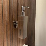 Le distributeur de savon Sonego Chrome de yessential.fr est un distributeur mural moderne en acier inoxydable avec couvercle à pompe et fenêtre pour afficher le niveau de savon, monté sur un panneau de salle de bain en bois.
