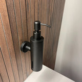 Le distributeur de savon mural Alberto Black de yessential.fr en noir mat est fixé à un bâton en bois au-dessus d'un lavabo de salle de bain blanc.