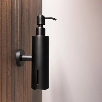 Le distributeur de savon Alberto Black de yessential.fr est fixé au mur sur un panneau en bois, mettant en valeur son design moderne et épuré. Le côté droit de l’image montre un arrière-plan uni aux tons clairs.