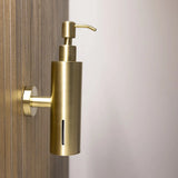 La pompe à savon Alberto Gold de yessential.fr avec support mural en acier inoxydable et fente de fenêtre verticale se fixe sur un mur en bois, sur un fond neutre.